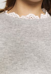 Col en dentelle œillet blanche sous un pull en tricot gris clair sur une personne aux longs cheveux bruns.
