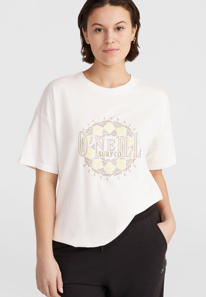White cotton t-shirt with oversized fit, featuring a rounded hem and a central graphic logo in beige and yellow. Paired with black pants.