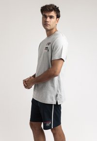 Camiseta de algodón gris con mangas cortas, con un diseño de texto en rojo y azul. Combinada con pantalones cortos azul marino que muestran un logotipo y un pespunte de contraste.