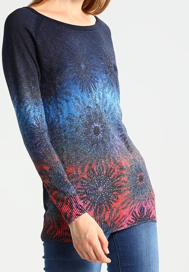 Femme portant un pull à manches longues avec des motifs circulaires en forme d'explosion d'étoiles bleues, violettes et rouges, associée à un jean bleu, debout devant un fond uni.