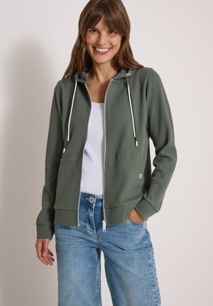 Femme souriante portant un sweat à capuche zippé vert olive sur un haut blanc et un jean bleu clair, une main dans la poche, sur un fond uni.