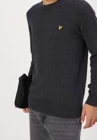Persona vestida con un suéter gris oscuro con un logo de pájaro amarillo en el pecho, jeans grises y sosteniendo un pouch negro con una correa de marca.