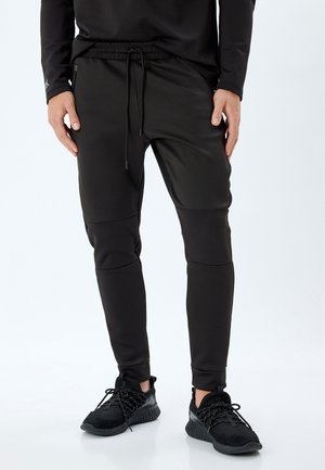 Zwarte sportieve joggers met een verstelbare tailleband, tapered fit, kniestukken en een zachte, gestructureerde stof. Gecombineerd met zwarte sportschoenen.