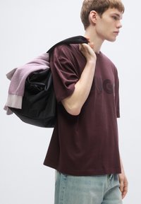 Mørk bordeaux oversized t-shirt med et hævet logo, kombineret med lyseblå jeans. Sort taske med grå accenter og en lavendelfarvet sweater lagt over.