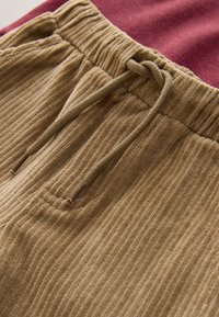 Pantalon en velours côtelé de coton beige avec une texture côtelée verticale. Taille élastique avec un cordon de serrage. En arrière-plan, un tissu bordeaux.