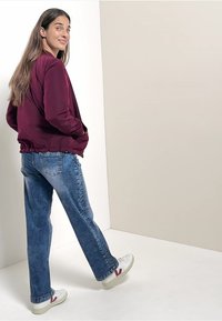 Bordowa kurtka bomberka z prążkowanymi mankietami, w połączeniu z jasnoniebieskimi spodniami o wysokim stanie i białymi trampkami z czerwonymi akcentami.