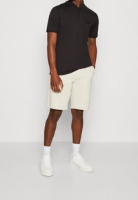 Schwarzes Polohemd mit kurzen Ärmeln, zwei Knöpfen und einem kleinen Logo. Kombiniert mit cremefarbenen Shorts und weißen Sneakers.