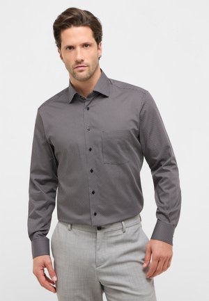 Graues Langarmshirt mit kleinem weißen Punktmuster, Button-Down-Kragen, schwarzen Knöpfen und einer einzelnen Brusttasche, getragen mit hellgrauer Hose.
