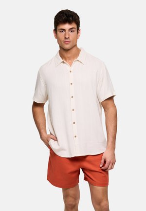 Hombre con camisa beige de manga corta abotonada y pantalones cortos de color naranja quemado, de pie con una mano en el bolsillo contra un fondo liso.