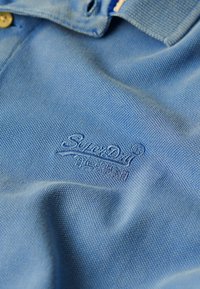 Polo in tessuto azzurro chiaro con trama testurizzata, dotato di un logo ricamato in filo più scuro sul lato sinistro. Dettaglio dei bottoni visibile.