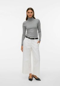 Mujer de pie con una mano en el bolsillo, vistiendo un suéter gris de cuello alto, pantalones blancos de pierna ancha con cinturón negro y tacones negros de punta puntiaguda.