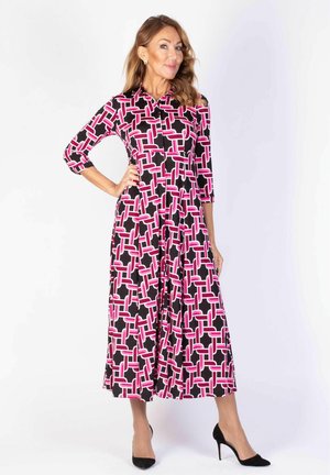 Nainen mustassa, vaaleanpunaisessa ja valkoisessa kuvioidussa pitkähihanaisessa mekossa, joka seisoo yhdellä kädellä lantiolla ja pukeutuneena mustiin korkokenkiin.