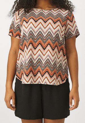 Kvinde iført en kortærmet zigzag-mønstret top i orange, sort og hvid med ensfarvede sorte shorts.