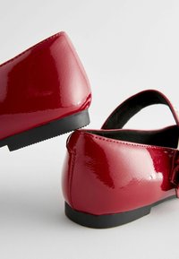 Rote Lacklederschuhe mit einer glatten, glänzenden Oberfläche, runden Zehen und einer niedrigen schwarzen Gummisohle. Es gibt einen Riemen über dem Spann.