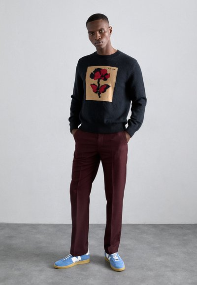 Μαύρο πλεκτό πουλόβερ με λουλουδάτο σχέδιο σε κόκκινο και μαύρο σε μπεζ φόντο, συνδυασμένο με βουργουνδί παντελόνι και μπλε αθλητικά παπούτσια.