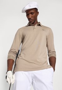 Bēšas krāsas garām piedurknēm polo krekls ar trīs pogu apkakli un ribotām aprocēm, apvienots ar baltām biksēm. Fona attēlā redzama balta cepure un golfa nūja.