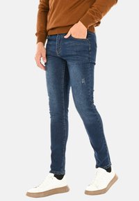 Jeans skinny blu scuro in denim, con un delicato effetto strappato, dotati di tasche frontali, abbinati a sneakers bianche e a un maglione marrone.
