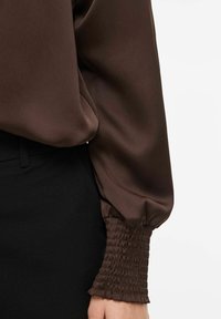 Brūna satīna blūze ar savāktām rievotām aprocēm, gludu tekstūru un brīvu piegriezumu, apvienota ar melnu audumu vidukļa zonā.