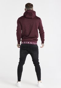 Uomo con tatuaggi che indossa una felpa con cappuccio bordeaux maculata e jeans skinny neri, in piedi di spalle su uno sfondo bianco.