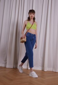 Un crop top vert à côtes, un jean bleu taille haute et des baskets blanches. Un sac en bandoulière tissé beige ajoute de la texture. L'arrière-plan présente des rideaux doux.