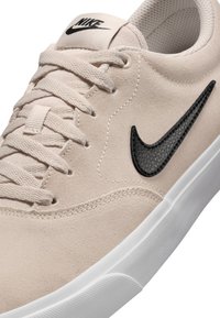 Zapatilla de ante beige con un distintivo negro de Nike texturizado, cordones redondos y suela de goma blanca. Diseño suave y minimalista.