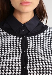 Kvinna klädd i en svart knappad skjorta med krage under en svart och vit houndstooth-mönstrad cardigan med svarta knappar.