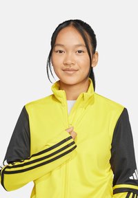 Gelbe Zip-Jacke mit schwarzen Ärmeln, die drei schwarze Streifen auf jedem Ärmel hat. Hergestellt aus leichtem, glattem Stoff.