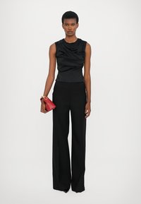 Black sleeveless top with gathered texture, paired with black wide-leg trousers. Model holds a small red handbag with gold hardware.