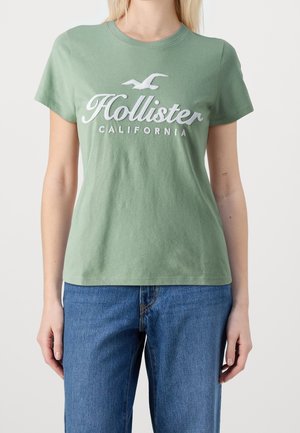 Green cotton t-shirt with short sleeves. Features white embroidered "Hollister California" text and subtle bird logo. Classic crew neck design.