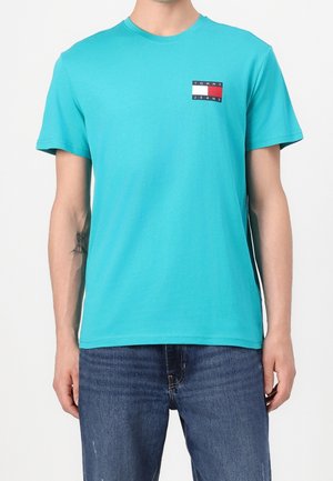 Mężczyzna w turkusowej koszulce z krótkim rękawem z małym logo Tommy Jeans na lewej piersi i niebieskich dżinsach.