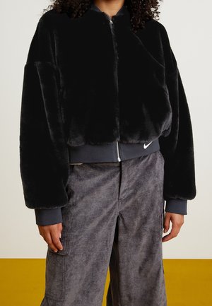 Osoba nosząca czarną kurtkę ze sztucznego futra na zamek błyskawiczny, z ściągaczami na rękawach i w talii, w połączeniu z luźnymi szarymi spodniami typu cargo.