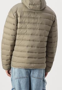 Khaki prošívaná bunda s kapucí, horizontální prošívání, měkká textura a elastické manžety, nošená přes světle modré džínové kalhoty.