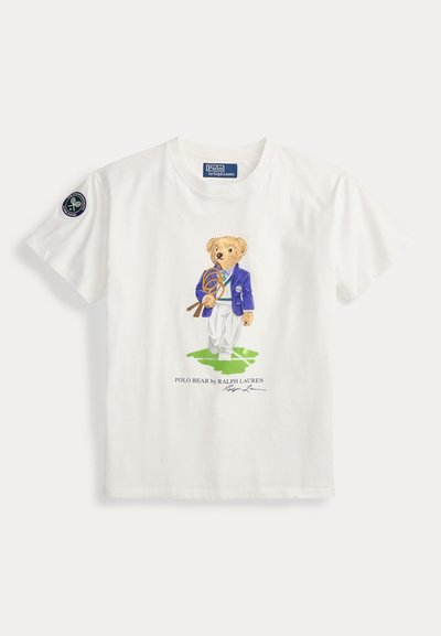 T-shirt blanc à manches courtes avec un ours en peluche en blazer bleu tenant des raquettes de tennis debout sur l'herbe, texte Polo Bear de Ralph Lauren en dessous.