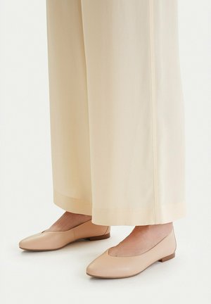 Beżowe płaskie buty noszone z kremowymi spodniami o szerokich nogawkach, prezentujące stopy i dolne partie nóg na jednolitym tle.