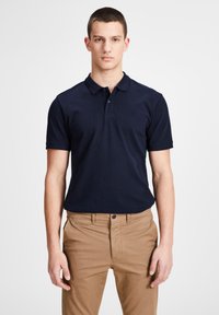 Polo shirt bleu marine avec col, manches courtes et un logo discret. Porté avec un pantalon kaki, présentant un bouton et un détail de poche.