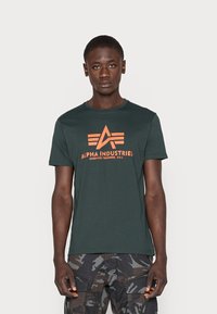 Tricou din bumbac verde închis, cu un logo Alpha Industries portocaliu, având un design cu stea și dungi, mâneci scurte și un guler clasic de tip crew.