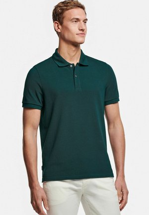 Man draagt een donkergroen poloshirt met korte mouwen en gebroken witte broek, kijkt zijwaarts tegen een witte achtergrond.