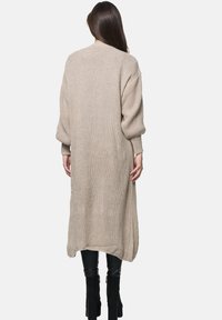 Lång, beige stickad cardigan med ribbad textur, översized ärmar och sänkt nederkant. Bärs över svarta tighta byxor och svarta stövlar.
