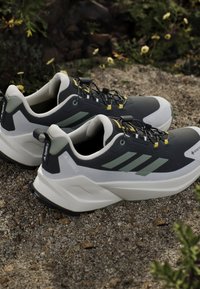 Para czarno-białych butów trekingowych Adidas Terrex z zielonymi paskami odpoczywająca na żwirze na zewnątrz w pobliżu roślin.