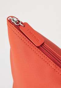 Trousse rouge texturée avec fermeture éclair, dotée d'un curseur assorti rouge et d'une étiquette logo en relief, fabriquée en matériau synthétique durable.