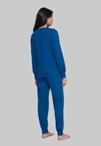Felpa blu e pantaloni coordinati in un materiale morbido. Maniche lunghe e polsini. Design semplice e privo di decorazioni; indossata a piedi nudi. Vista da dietro.
