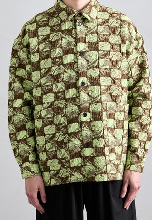 Mężczyzna ubrany w koszulę na guziki z długim rękawem o wzorze z brązowych i jasnozielonych teksturowanych kwadratów, w połączeniu z czarnymi spodniami, stoi na jednolitym tle.