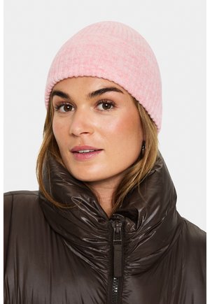 Žena se šedým pleteným čepicím a lesklou tmavohnědou puffer bundou s vysokým límcem na jednobarevném pozadí.