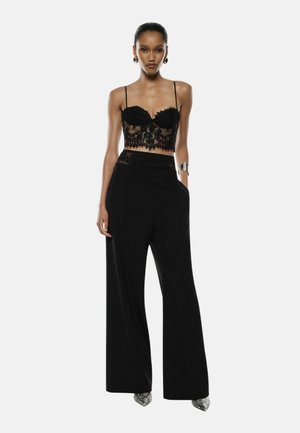 Donna con un crop top in pizzo nero a maniche sottili, pantaloni neri a vita alta e gamba larga, bracciale in argento e scarpe a punta in pelle di serpente.