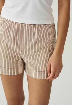 Žena nosi béžové a biele šortky s vertikálnymi pruhmi a biele tričko, stojaca s pravou rukou voľne pri boku.