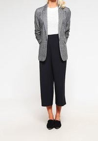 Blazer gris texturé sur un top en maille blanche, associé à un pantalon noir raccourci et des ballerines noires. Lignes simples et silhouette ajustée.