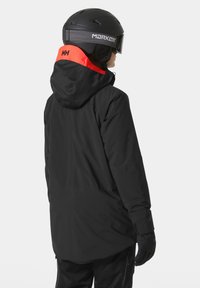 Czarna kurtka narciarska z czerwonym akcentem na kapturze, o luźnym kroju, wykonana z wodoodpornego materiału i z regulowanymi mankietami. W komplecie z czarnym kaskiem.
