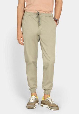 Hombre vistiendo pantalones jogger beige con puños elásticos y cintura con cordón, combinado con zapatillas beige y amarillas, de pie contra un fondo blanco.