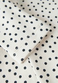 Bavlněná látka s žebrovanou texturou, s krémovým pozadím a rovnoměrně rozmístěnými černými puntíky. Převážně záhyby ukazující detail vzoru.