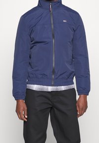 Granatowa kurtka na suwak z kołnierzem typu stójka, elastycznymi mankietami i dołem. Na lewej piersi małe logo oraz kratkowaną podszewkę na dole.